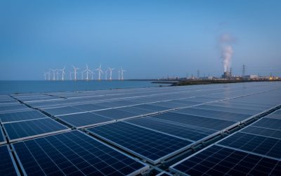 Wide view of a modern industrial facility with solar panels in the foreground, offshore wind turbines in the distance, and a faint geothermal steam plume rising behind.