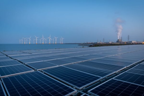 Wide view of a modern industrial facility with solar panels in the foreground, offshore wind turbines in the distance, and a faint geothermal steam plume rising behind.